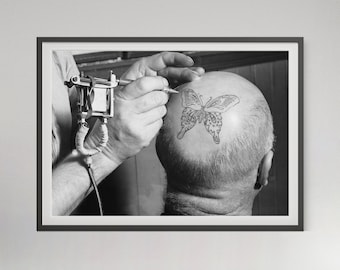May include: Black and white framed photograph of a person's head with a butterfly tattoo being applied. A tattoo artist is using a tattoo machine to create the butterfly design on the person's head. The image is a close-up shot.