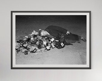 May include: A black and white photograph featuring a person wearing a dark coat and cap lying on the ground, resting their head on a large pile of various glass bottles. A vintage box camera is positioned directly next to the person on the ground.