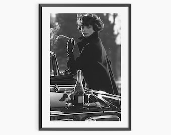 May include: A black and white framed photograph featuring a person in a dark coat and gloves leaning against a vintage car. A champagne bottle and two glasses sit on the car hood. The person holds a smoking cigarette, looking toward the camera.