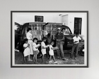May include: A black and white photograph of a mobile library van with open side panels revealing shelves filled with books. Several individuals stand around the vehicle, some holding books, while a driver sits in the front seat of the car.