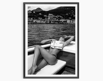 May include: A black and white framed photograph depicting a person reclining on a boat seat with a newspaper covering their upper body. The background features a body of water and a coastal town nestled against a mountain range under a cloudy sky.