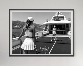 May include: A black and white framed wall art print depicting two individuals in white athletic wear and caps playing tennis on a court located on the deck of a large yacht. The court surface features a white anchor logo and the text YACHT CLUB.