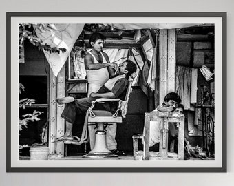 May include: A black and white photo of a barber shop in India. A man sits in a barber chair getting a shave from another man. A third man sits in a chair nearby, watching.
