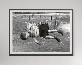 May include: A black and white framed photograph depicting a child wearing a striped shirt and dark shorts hanging from a clothesline by their waistband with wooden clothespins. The child is suspended horizontally above a field with dark shoes.