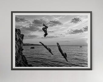 May include: A black and white framed wall art print depicting three figures diving from a rocky cliff into the ocean at sunset. The composition captures one person mid-flip and two others in a downward dive toward the water under a cloudy sky.