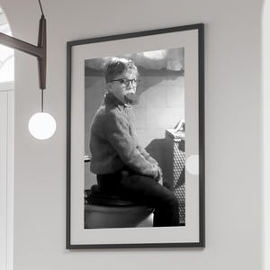 May include: Monochrome framed print featuring a young person in glasses and a sweater, holding a lollipop. The image is a vintage photograph, with the subject seated on a toilet. The frame is black, and the print is displayed on a white wall.