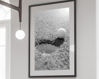 Op de afbeelding: Een zwart-witfoto van een golfbal die op een groene putting green ligt, in de buurt van een golfhole.