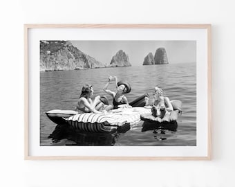 May include: A black and white framed photograph depicting three individuals on inflatable rafts in the ocean. They are gathered around a floating table with drinks and snacks, with large rock formations visible in the background of the sea.