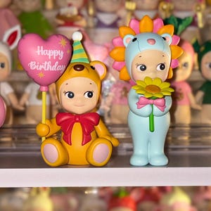 May include: Two small collectible figurines. One is a yellow bear wearing a party hat and holding a pink heart-shaped balloon with the text Happy Birthday. The other is a light blue figure wearing a lion mane hood and holding a yellow flower.