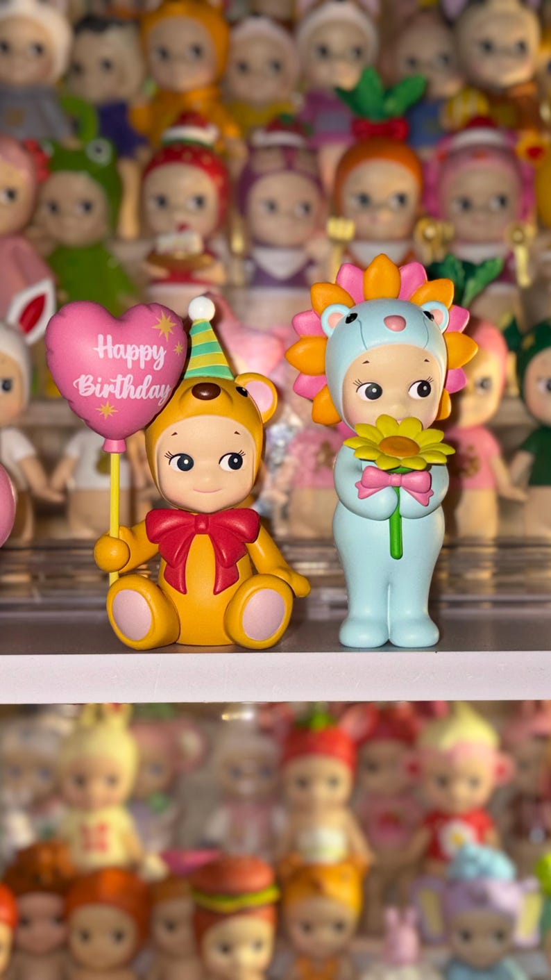 May include: Two small collectible figurines. One is a yellow bear wearing a party hat and holding a pink heart-shaped balloon with the text Happy Birthday. The other is a light blue figure wearing a lion mane hood and holding a yellow flower.