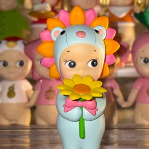 May include: A small figurine of a character wearing a light blue lion costume with a bright orange and pink mane. The character holds a yellow flower with a green stem and has a small pink bow at the base of the flower, standing upright.