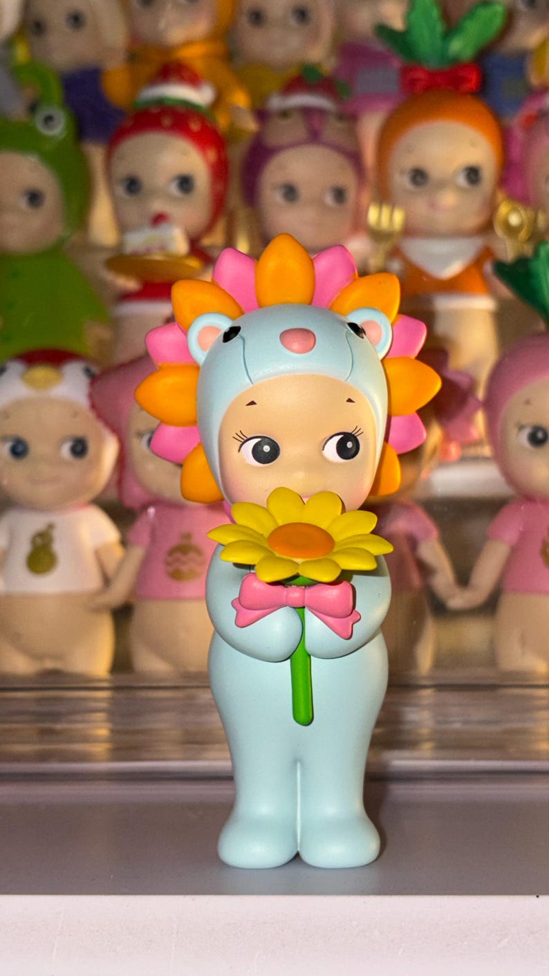 May include: A small figurine of a character wearing a light blue lion costume with a bright orange and pink mane. The character holds a yellow flower with a green stem and has a small pink bow at the base of the flower, standing upright.