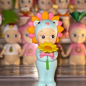 May include: A small figurine of a character wearing a light blue lion costume with a bright orange and pink mane. The character has a pale complexion, large dark eyes, and is holding a yellow flower with a green stem and a small pink bow.