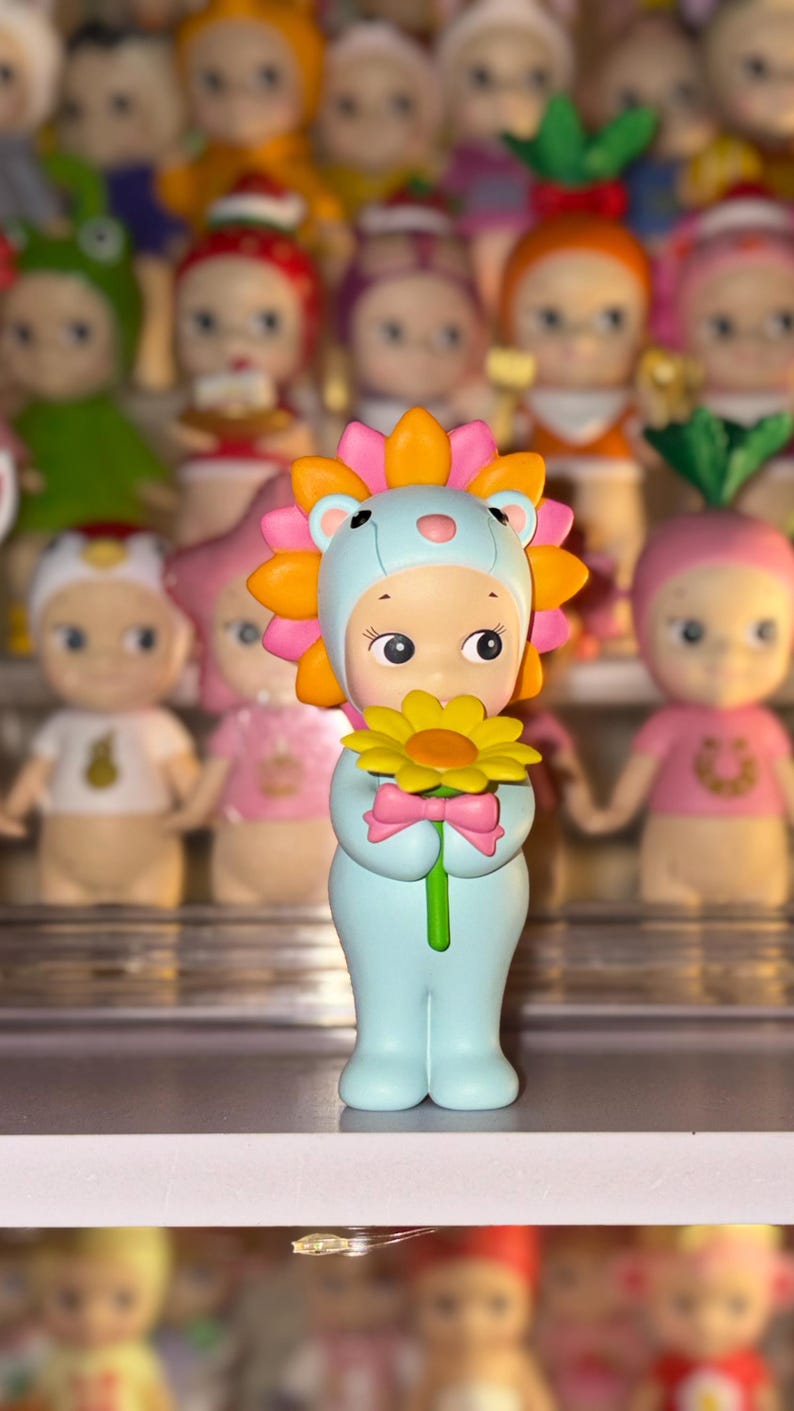 May include: A small figurine of a character wearing a light blue lion costume with a bright orange and pink mane. The character has a pale complexion, large dark eyes, and is holding a yellow flower with a green stem and a small pink bow.