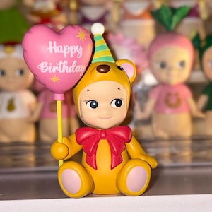 May include: A yellow bear figurine wearing a party hat and a red bow, holding a pink heart-shaped balloon with the text Happy Birthday printed in white. The bear has light skin, blue eyes, and pink accents on its ears, hands, and feet.