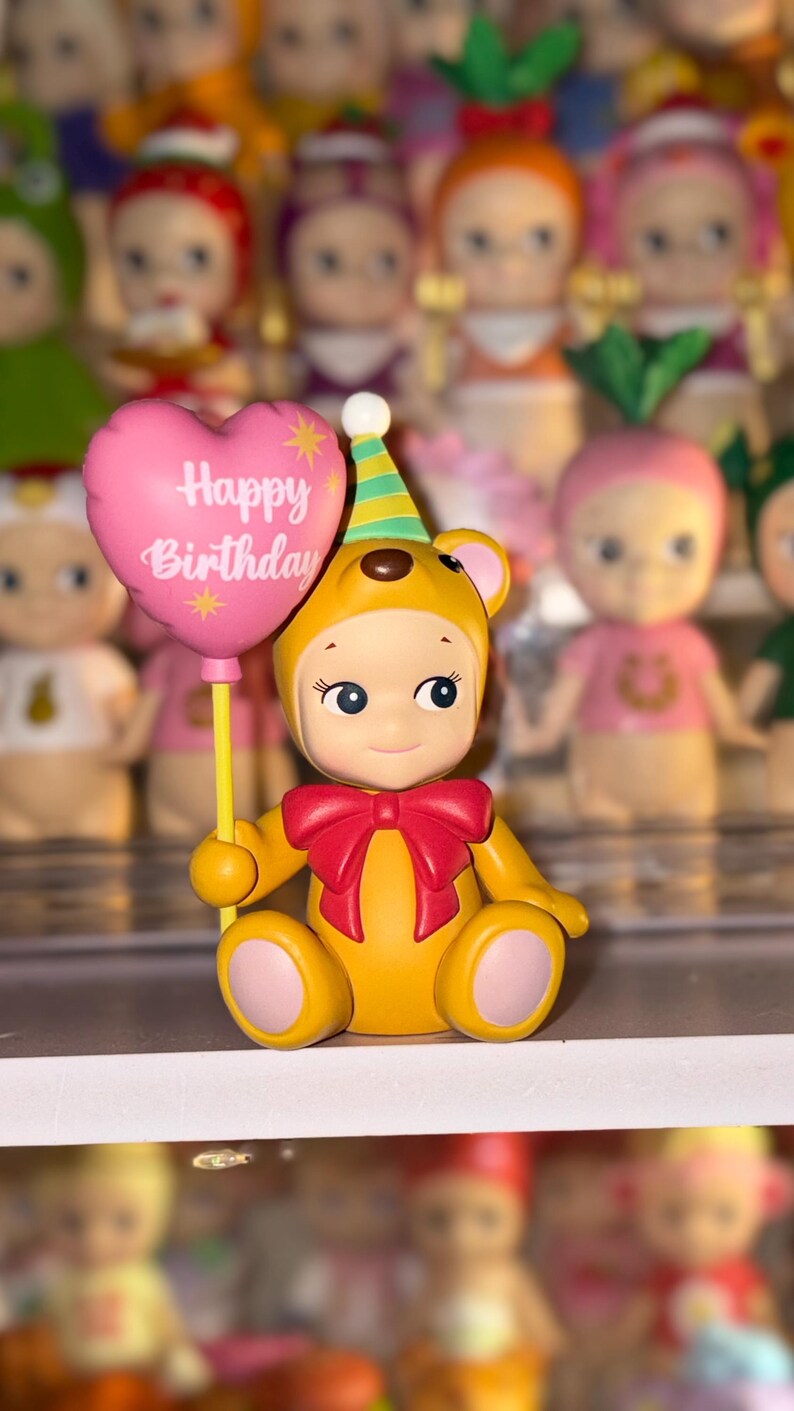 May include: A yellow bear figurine wearing a party hat and a red bow, holding a pink heart-shaped balloon with the text Happy Birthday printed in white. The bear has light skin, blue eyes, and pink accents on its ears, hands, and feet.