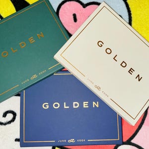 Puede incluir: Tres libros de tapa dura con la palabra "GOLDEN" en letras doradas. Los libros son de color verde azulado, blanco y azul marino. El texto "JUNG ♾️ KOOK" está impreso en la parte inferior de cada libro.