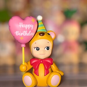 May include: A yellow bear figurine wearing a green and yellow striped party hat and a red bow tie. The bear holds a pink heart-shaped balloon with the text Happy Birthday printed in white, featuring small yellow star accents on the balloon surface.
