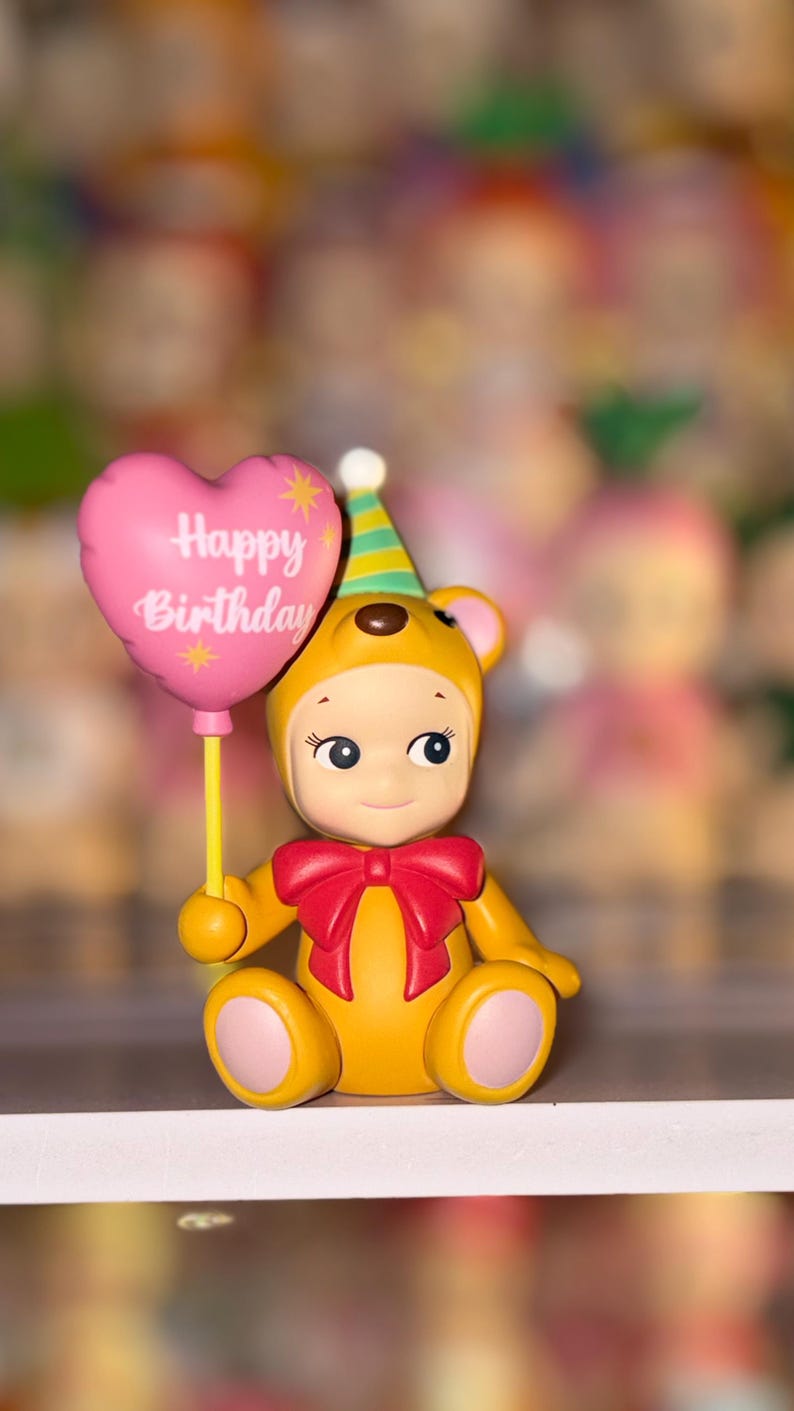 May include: A yellow bear figurine wearing a green and yellow striped party hat and a red bow tie. The bear holds a pink heart-shaped balloon with the text Happy Birthday printed in white, featuring small yellow star accents on the balloon surface.