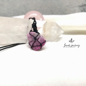 Puede incluir: Un colgante de cristal rosa envuelto en un cordón negro, colgando de un collar de cordón negro. El cristal tiene una forma rugosa y natural y es de color rosa y blanco.