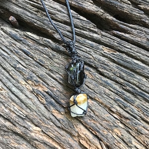 Puede incluir: Un collar hecho a mano con dos piedras preciosas naturales. La piedra superior es un rectángulo oscuro y translúcido, y la piedra inferior tiene una forma irregular de color amarillo claro. Ambas están sujetas por un cordón negro tejido, suspendido de un cordón a juego.