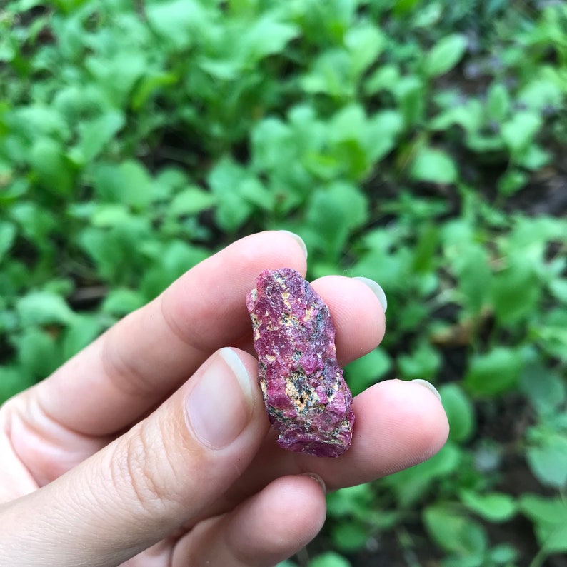 Op de afbeelding: Een close-up van een roze en rode edelsteen met een ruwe, natuurlijke textuur. De edelsteen wordt in een hand gehouden.