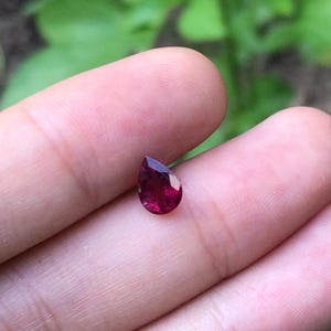 May include: A pear-shaped, deep red gemstone. The gem is held between fingers, with a blurred green background. The stone's facets catch the light, showcasing its rich color and clarity. This gemstone is likely a ruby.
