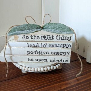 May include: A stack of four white rectangular wooden blocks with black text reading do the right thing, lead by example, positive energy, and be open minded. The stack is topped with two green fabric leaves and twine, resting on a white beaded riser.