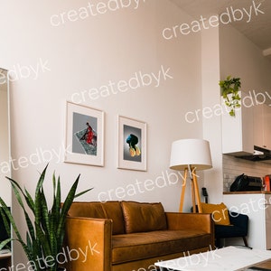 May include: A living room interior with a brown leather sofa, a white coffee table, a potted snake plant, and two framed art prints on the wall. The room has white walls and a white ceiling fan.