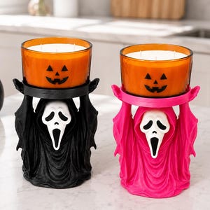 May include: Two candle holders shaped like hooded figures with white ghost masks, one in black and one in bright pink. Each holder supports a round orange glass candle jar featuring a black jack-o'-lantern face print with white wax inside.