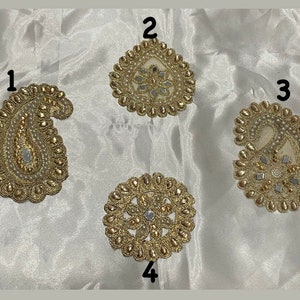 May include: Four gold and white embroidered fabric patches with mirror accents. The patches are in the shape of a paisley and a circle.