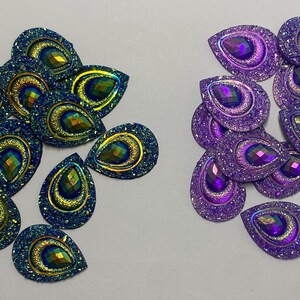 May include: Assortment of iridescent teardrop-shaped embellishments with glitter. The embellishments are in two colours: teal and purple.