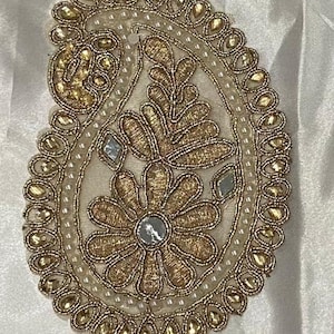 May include: A gold and white paisley-shaped embellishment with pearl and rhinestone accents. The embellishment is ideal for adding a touch of elegance to clothing, accessories, or home decor.