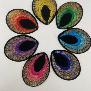 May include: Seven embroidered patches in the shape of an eye with a black centre and a gold outline. The patches are arranged in a circle and are in the colours red, yellow, green, blue, purple, pink, and blue.