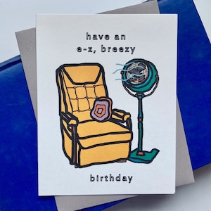 May include: A greeting card featuring a yellow armchair and a green fan. The text on the card reads "have an e-z, breezy birthday".