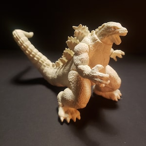 Könnte beinhalten: Hellbeige Godzilla-Figur in dynamischer Pose mit offenem Maul. Das Spielzeug hat detaillierte Schuppen, Rückenflossen und Krallen. Die Figur ist etwa 20 cm groß.