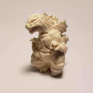 Puede incluir: Una figura de plástico blanca de Godzilla, un famoso monstruo de las películas japonesas. La figura está en posición de pie con los brazos levantados y la boca abierta en un rugido.