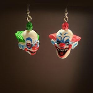 Könnte beinhalten: Ein Paar Ohrringe mit gruseligen Clown-Gesichtern. Der Clown links hat grünes Haar und ein weißes Gesicht mit blauen und roten Details. Der Clown rechts hat rotes Haar und ein weißes Gesicht mit blauen und roten Details.