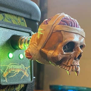 Peut inclure: Une sculpture détaillée d'un crâne avec de la matière cérébrale exposée, enveloppée de bandages beiges. Le crâne est attaché à une machine verte et noire avec le texte "Metallica" visible. La sculpture a un thème d'horreur.