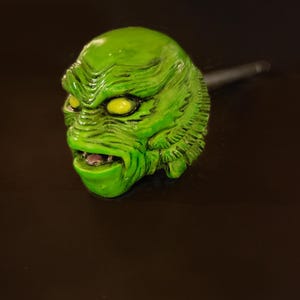 Op de afbeelding: Groen schepsel hoofd met gele ogen en een tandenrij. Het schepsel is gemaakt van plastic en heeft een textuur.