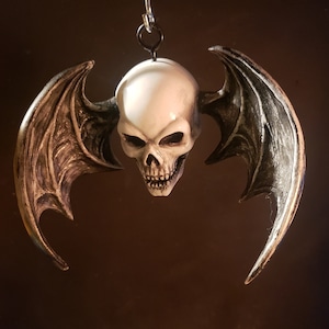 May include: A white skull with black wings hanging from a silver chain. The skull has a detailed face with a menacing expression. The wings are detailed with textured feathers.