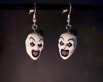 Könnte beinhalten: Ein Paar Ohrhänger mit Miniatur-Clownsköpfen in Weiß, die schwarze Zylinder, schwarzes Augen-Make-up und weit geöffnete Münder mit sichtbaren Zähnen aufweisen. Die Clownsköpfe sind an silberfarbenen Ohrhaken für durchstochene Ohren befestigt.