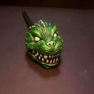 Puede incluir: Pípa de cerámica verde y blanca con una cara de monstruo detallada. El monstruo tiene dientes afilados y una boca abierta.