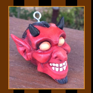May include: Red devil ornament with horns, a toothy grin, and glowing eyes. The ornament is made of clay and has a loop for hanging.