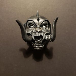May include: A detailed, metallic sculpture of a demonic face with horns, a nose ring, and a spiked crown. The face has an aggressive expression with sharp teeth and glaring eyes. The sculpture is dark grey with highlights.
