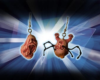 May include: A pair of unusual earrings with disturbing face-like designs. One earring is a fleshy, open-mouthed face, the other has a spider-like body with a face. Both are suspended from silver-coloured hooks.