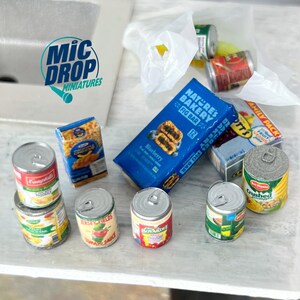 May include: A miniature grocery shopping scene with various canned goods, a box of pasta, a box of fig bars, and a bag of fruit. The cans include brands like Campbell's, Jersey Fields, and Boyardee. The scene is set on a light gray surface.