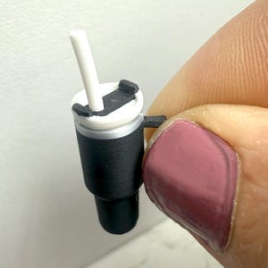 May include: Miniature black and white tumbler with a white straw. The tumbler has a textured black sleeve and a white lid with a black closure. The tumbler is held between two fingers.