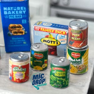 May include: A collection of miniature canned goods, including pork and beans, crushed pineapple, green beans, and corn. The cans are arranged on a table with a box of fig bars and a box of fruit juice in the background.