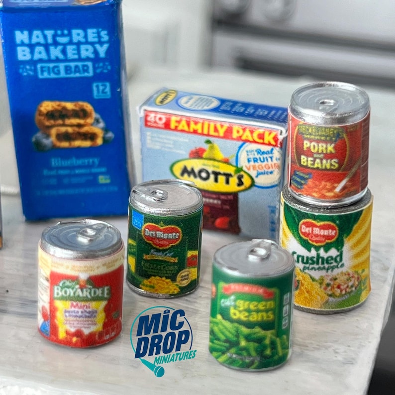 May include: A collection of miniature canned goods, including pork and beans, crushed pineapple, green beans, and corn. The cans are arranged on a table with a box of fig bars and a box of fruit juice in the background.